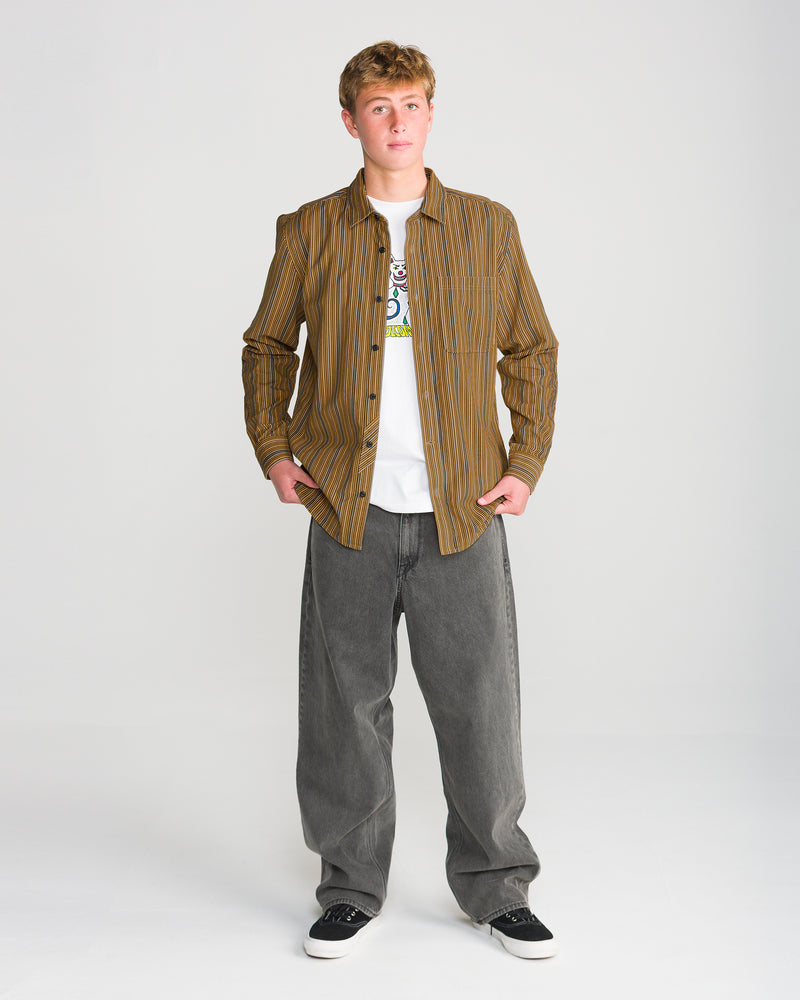 A person wearing a Volcom Men’s Featured Artist Casey Jones Long Sleeve unbuttoned over a white graphic t-shirt, paired with baggy grey jeans and black sneakers.