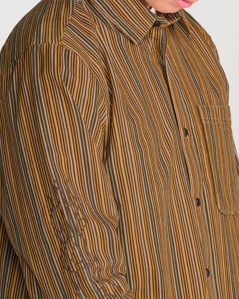 Close-up of a person wearing the Volcom Men’s Featured Artist Casey Jones Long Sleeve, a corduroy button-up shirt with vertical brown, tan, and teal stripes and a Casey Jones graphic embroidered on the right sleeve.