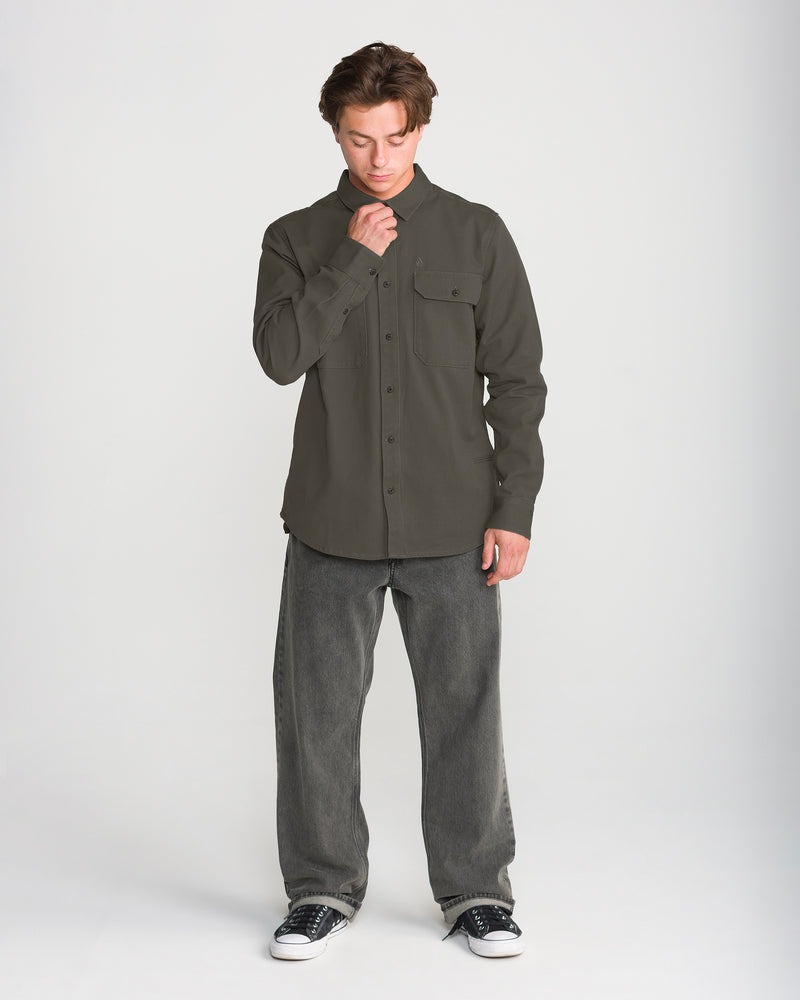 A man wearing a dark olive cotton twill Volcom Men’s Stockholm Long Sleeve with two chest pockets, relaxed-fit gray jeans, and black sneakers.