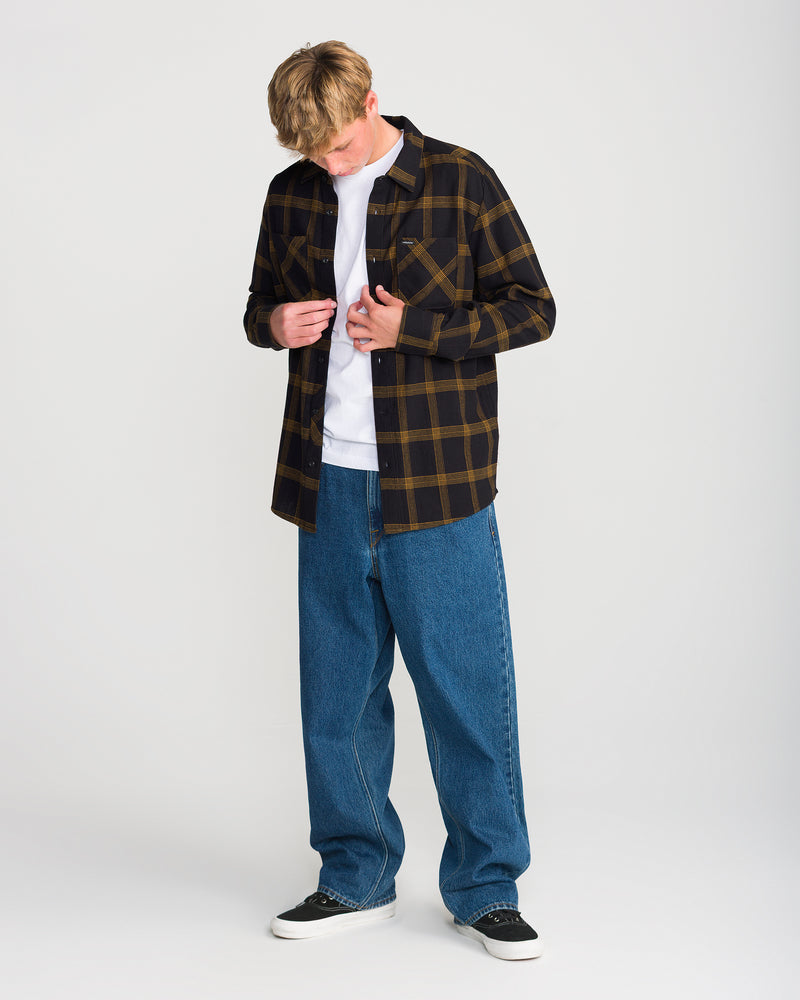 A person wearing an unbuttoned black Volcom Men’s Netastone Long Sleeve flannel shirt over a white t-shirt, baggy blue jeans, and black sneakers.