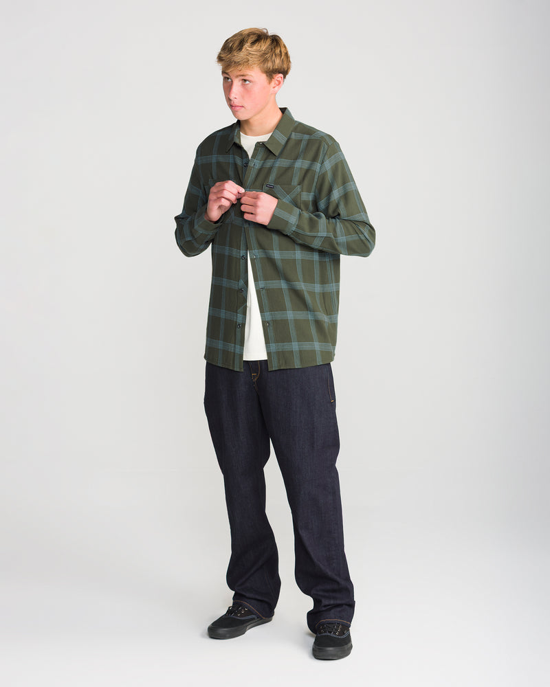 A person wearing a green plaid Volcom Men’s Netastone Long Sleeve over a white t-shirt, dark denim jeans, and black sneakers.