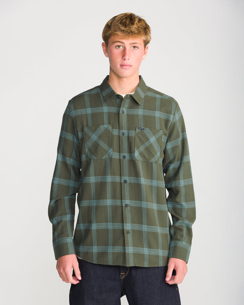 A man wearing a Volcom Men’s Netastone Long Sleeve, a classic fit, long-sleeve olive green and blue plaid flannel shirt with two chest pockets.