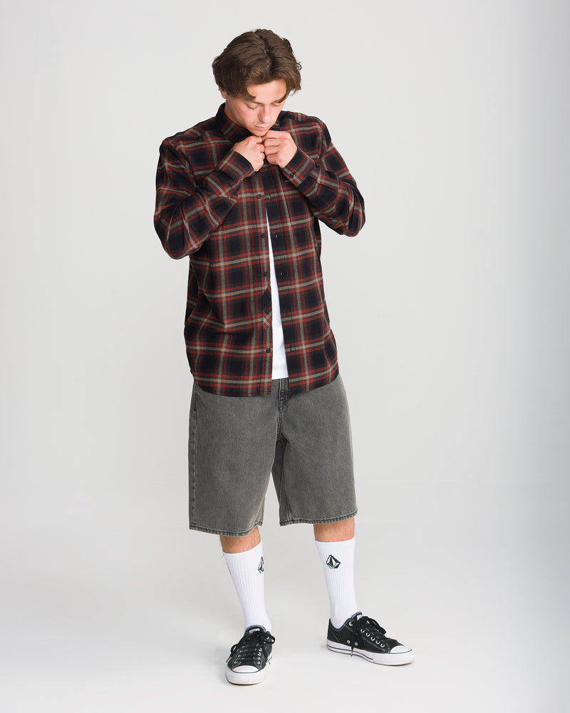 A man wears a red and black Volcom Men’s Caden Plaid Long Sleeve over a white t-shirt, gray denim shorts, white crew socks, and black low-top sneakers.