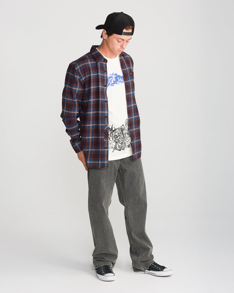 A person wearing a Volcom Men’s Caden Plaid Long Sleeve shirt over a graphic t-shirt, gray jeans, and black sneakers.