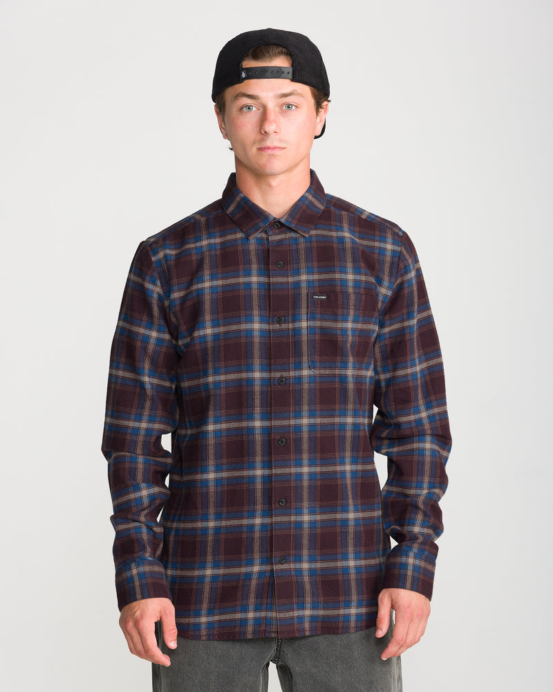 A man wears a Volcom Men’s Caden Plaid Long Sleeve brown and blue plaid yarn-dye cotton button-down shirt with a chest pocket and a black baseball cap.