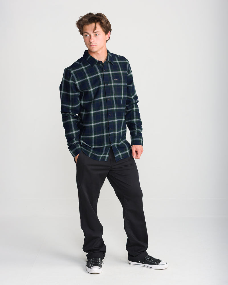 A man wearing a dark green and blue plaid Men’s Caden Plaid Long Sleeve Volcom cotton flannel shirt, black pants, and black sneakers.