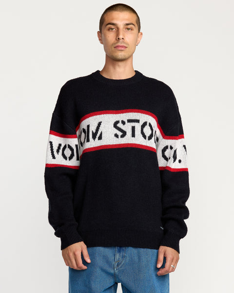 A man wears the Volcom Men's Carwash Sweater—black with red and white stripes and partial “VOM STO” text—standing against a plain light background.