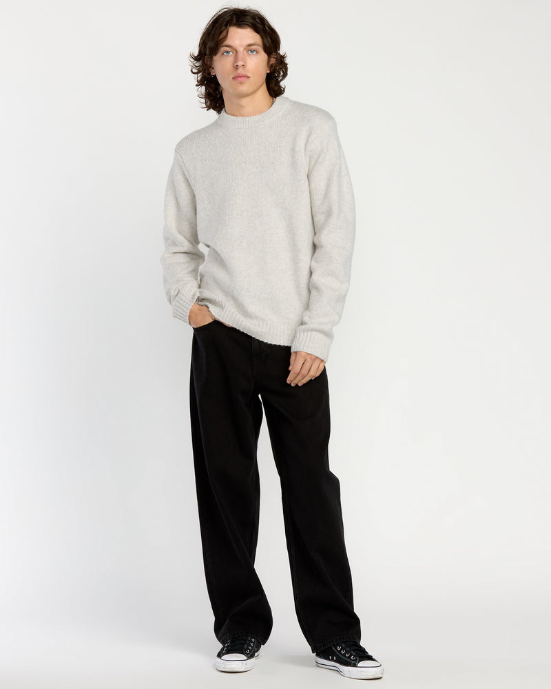 A person with medium-length brown hair wears the Volcom Men's Edmonder II Sweater in light gray, paired with loose black pants and black sneakers, standing against a plain white background with one hand in their pocket.