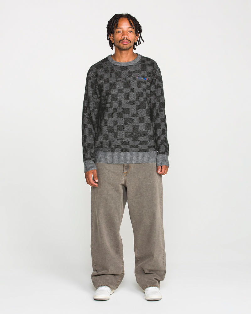 A person stands facing forward against a plain white background, wearing the Volcom Men's Featured Artist Antonio Carrau Sweater with a boxy fit, paired with loose brown pants and white sneakers.