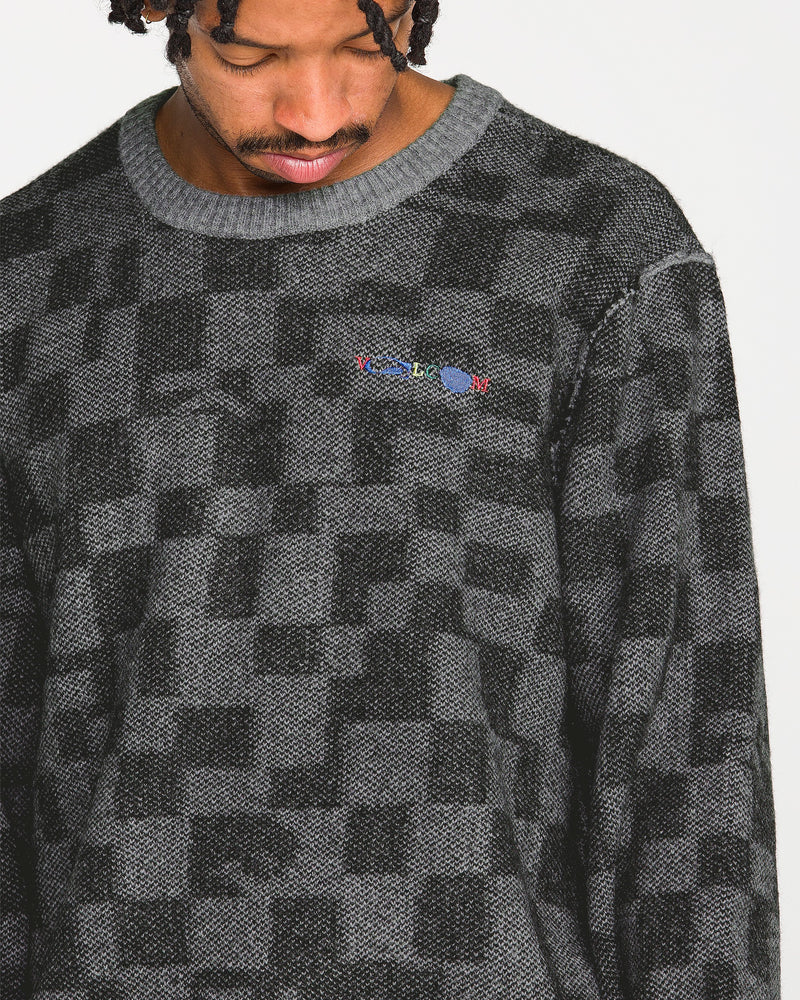 A person with short braids looks down, wearing the Volcom Men's Featured Artist Antonio Carrau Sweater, which features a textured gray and black checkered jacquard design with small, colorful embroidered text on the chest.