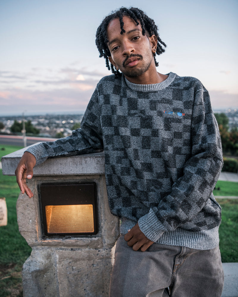 A man with short dreadlocks, dressed in the Volcom Men’s Featured Artist Antonio Carrau Sweater with a boxy fit, leans against a concrete structure under soft outdoor light at sunset, with a cityscape in the background.