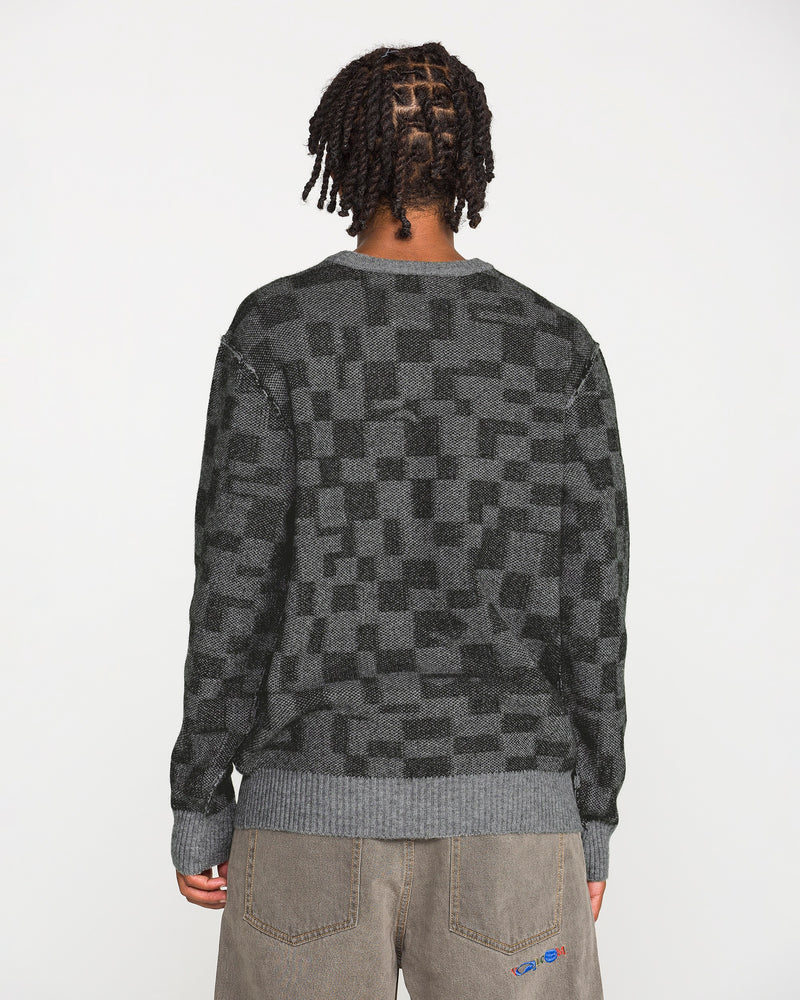 A person with short, twisted hair is seen from behind against a plain white background, wearing the Volcom Men's Featured Artist Antonio Carrau Sweater in dark gray checkered jacquard paired with brown pants.