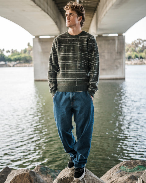 A person stands on a rock by the water under a bridge, wearing the Volcom Men’s Stone Optix Sweater with a crew neck and blue jeans, hands in pockets, looking to the side.