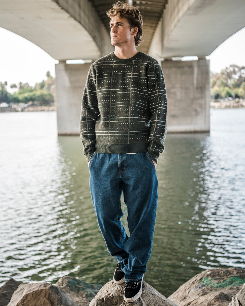 A person stands on a rock by the water under a bridge, wearing the Volcom Men’s Stone Optix Sweater with a crew neck and blue jeans, hands in pockets, looking to the side.