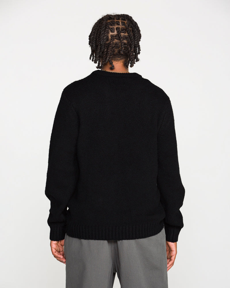 A person with braided hair stands facing away, wearing the Volcom Men's Edmonder II Sweater in black—crafted from recycled polyester—paired with gray pants, against a plain white background.