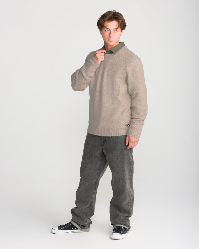 A young man wears the Volcom Men’s Edmonder II Sweater in a cashmere colorway, styled with a green shirt, gray jeans, and black sneakers. Standing against a plain white background, he looks to the side and adjusts his collar with one hand. Crafted from a recycled-poly blend for comfort and sustainability, his ensemble embodies effortless modern style.