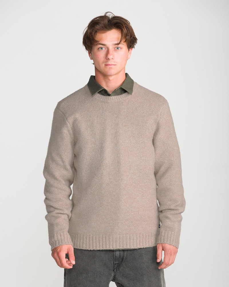 A young man with short brown hair wears the Volcom Men’s Edmonder II Sweater in a beige recycled-poly blend over a green collared shirt and dark pants. He stands facing forward against a plain, light background.
