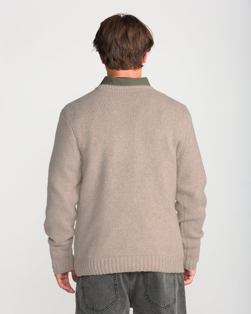 A person with short brown hair stands with their back to the camera, wearing the Volcom Men’s Edmonder II Sweater in the cashmere colorway—a cozy beige knit made from a recycled-poly blend—paired with gray jeans against a plain white background.