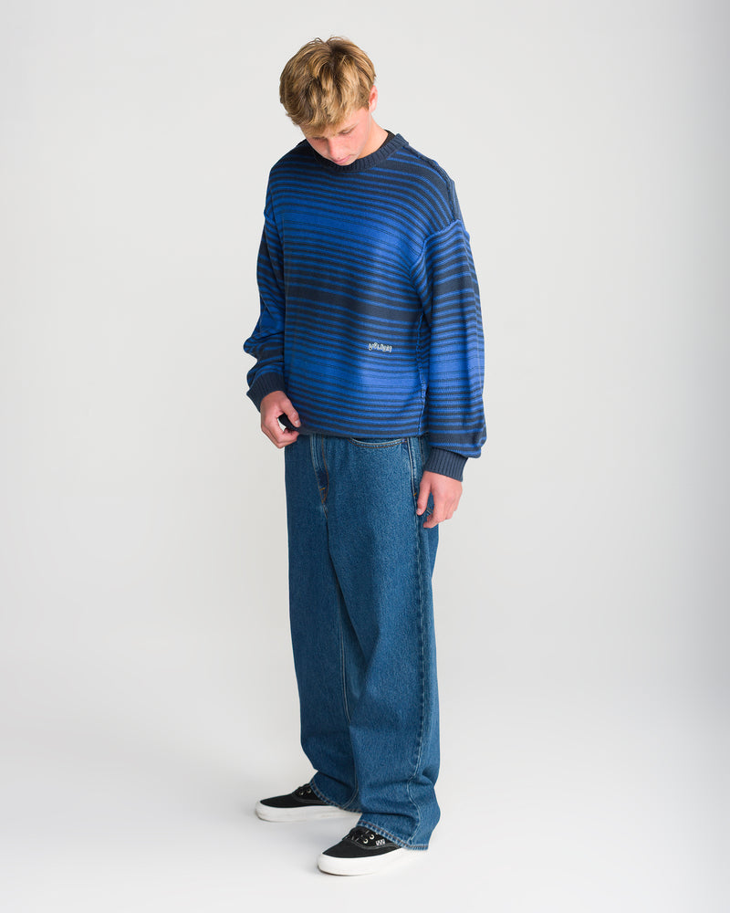 A person wearing a Volcom Men’s Fade In Crew Sweater in Nebulas Blue with black horizontal stripes, paired with loose-fit blue jeans and black sneakers.