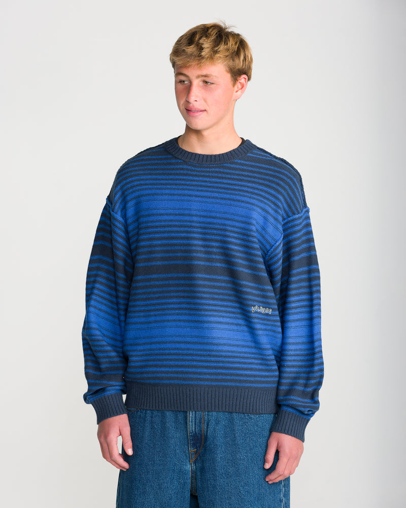 A person wearing a Volcom Men’s Fade In Crew Sweater, a Nebulas Blue wool-blend crewneck with blue and black horizontal stripes.