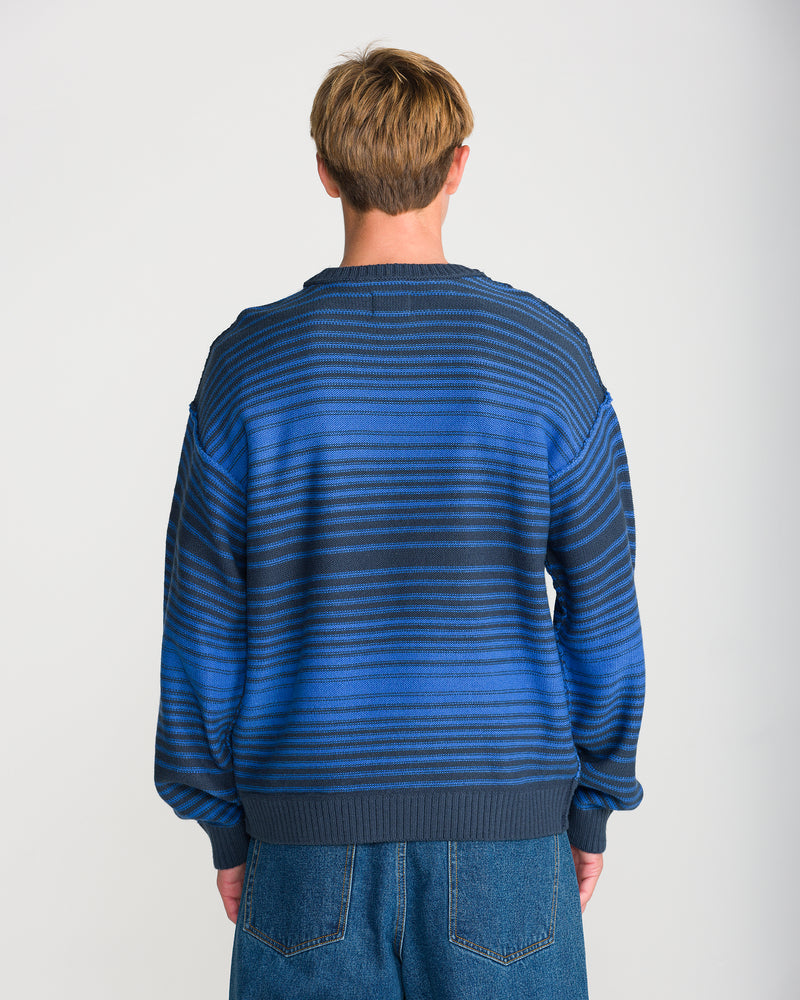 A person seen from behind, wearing a Volcom Men’s Fade In Crew Sweater in Nebulas Blue wool-blend with horizontal stripes and blue jeans.