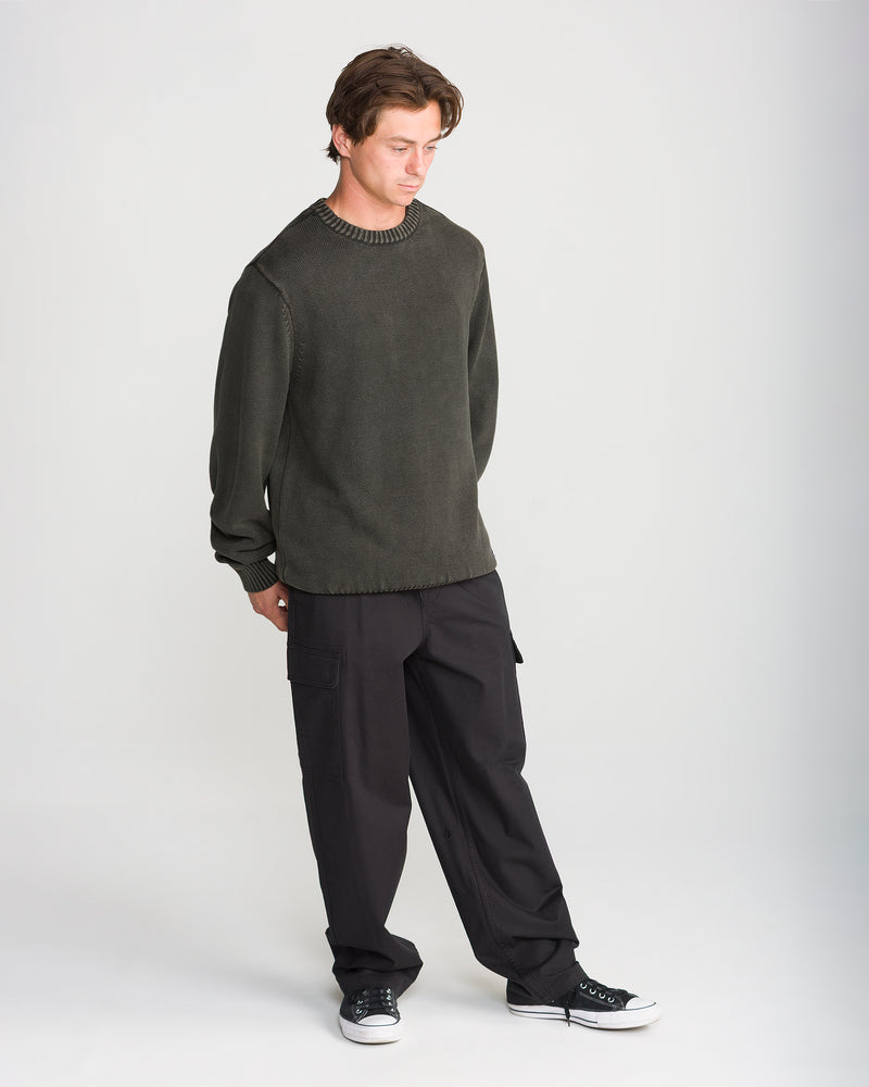 A man stands looking down wearing a dark green Volcom Men’s Bleached Crew Sweater, black cargo pants, and black low-top sneakers.
