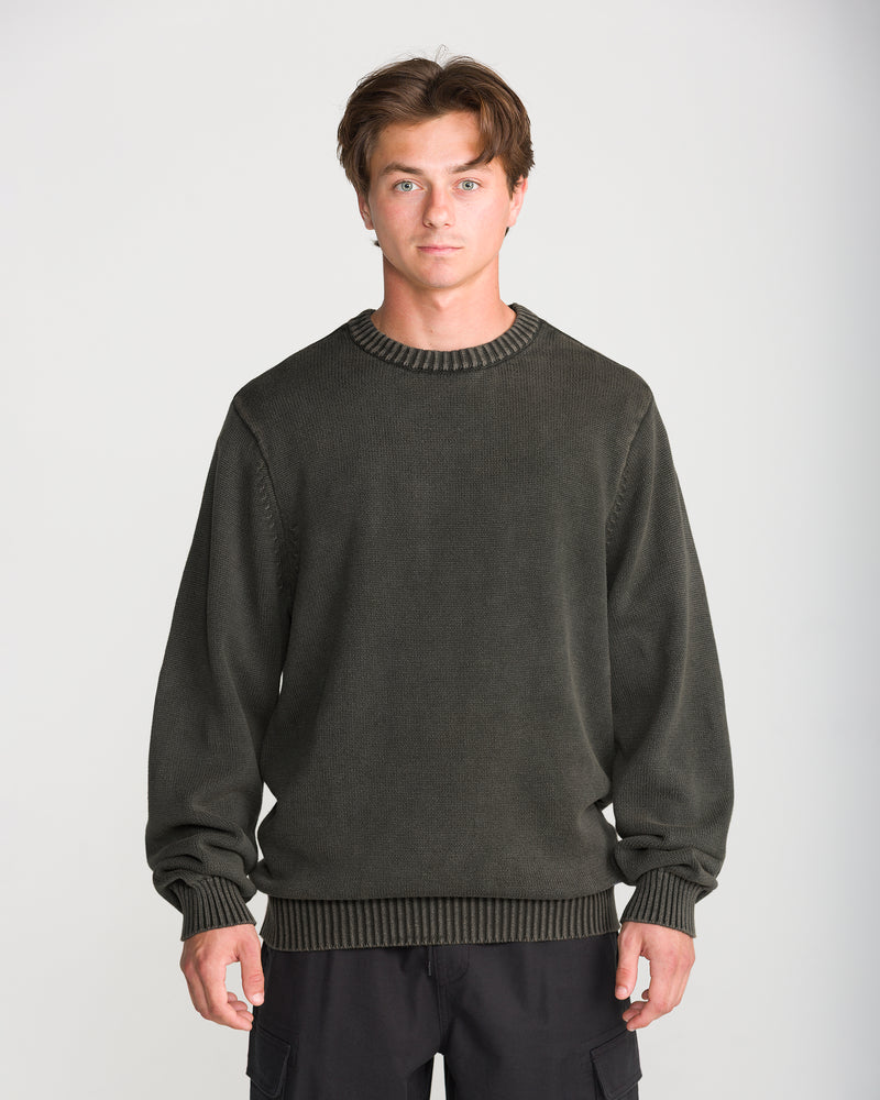 A man wearing a Volcom Men’s Bleached Crew Sweater in dark olive green with ribbed cuffs and hem.
