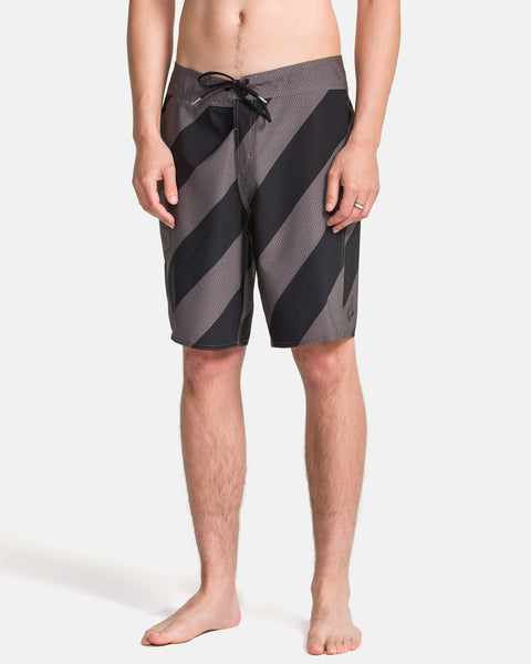 A man stands barefoot wearing Volcom Men's Manic Boardshorts with black and gray stripes and a drawstring waist, shown from the torso down against a plain white background.
