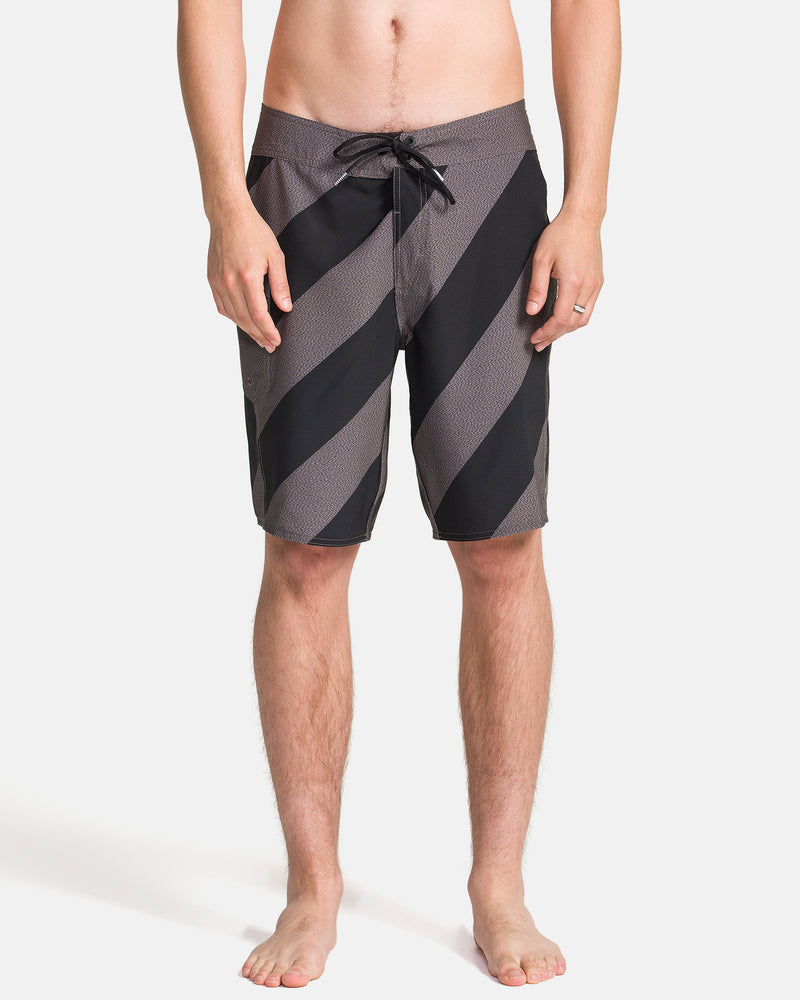 A person stands barefoot in Volcom Men's Manic Boardshorts, featuring knee-length black and gray diagonal stripes, shown from the front against a plain white background.