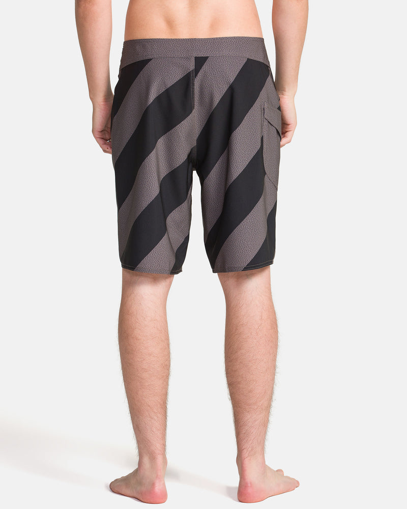A person stands barefoot facing away, wearing Volcom Men's Manic Boardshorts—black and gray with diagonal stripes and a back pocket—against a plain white background.