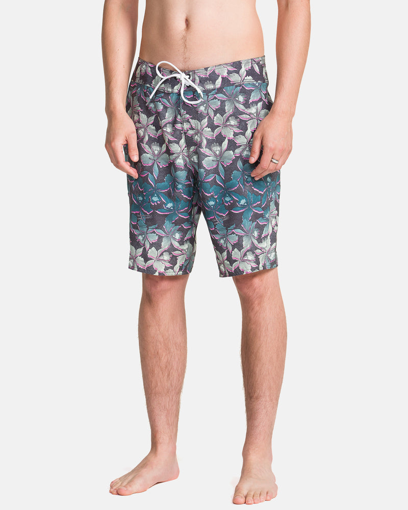 A man stands barefoot in Volcom Men's Manic Boardshorts featuring a tropical leaf pattern in green, purple, and teal with a white drawstring waist. Only his torso and legs are visible.