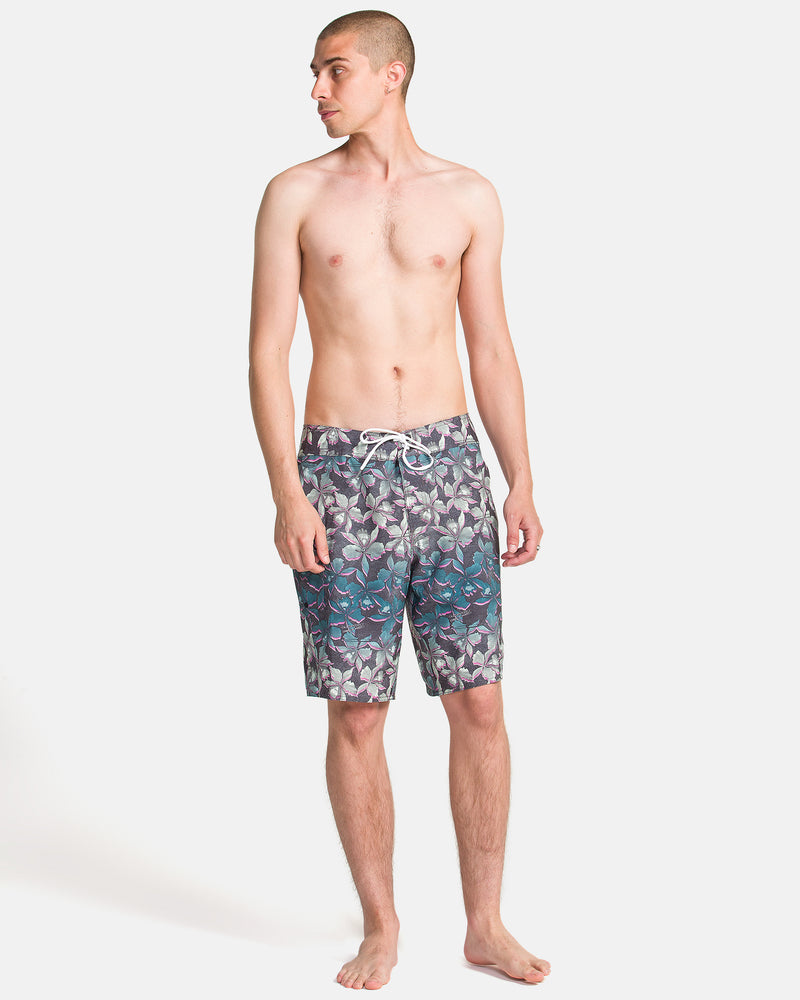 A barefoot man with a shaved head stands against a plain background, wearing Volcom Men's Manic Boardshorts with a floral pattern and drawstring, looking to his left.