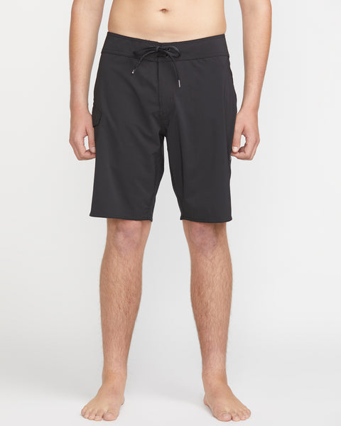 A person stands barefoot against a white background wearing Volcom Men's Lido Solid Mod 20 black knee-length swim trunks with a drawstring waist. Their hands are relaxed at their sides, and the upper body is partially out of frame.