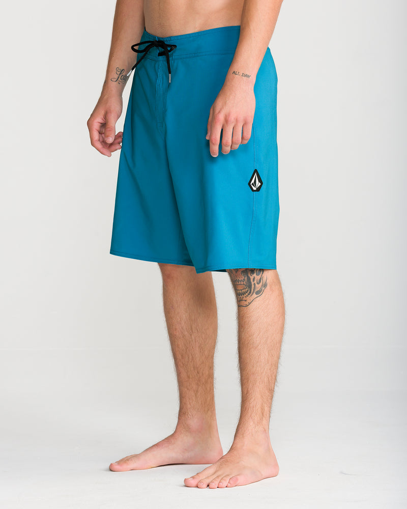 A man stands barefoot wearing the Volcom Men’s Lido Solid Mod 20 boardshorts in teal, featuring a black drawstring and a small logo on the left leg. His lower legs and arms are tattooed, and the background is plain and light-colored.