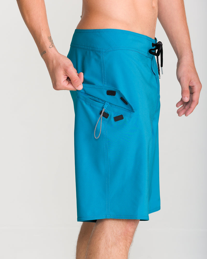 A person is seen from the waist down, against a plain background, wearing Volcom Men’s Lido Solid Mod 20 teal 4-way stretch boardshorts. Their right hand pulls open a side pocket featuring both a zipper and Velcro closure, revealing an interior key loop.