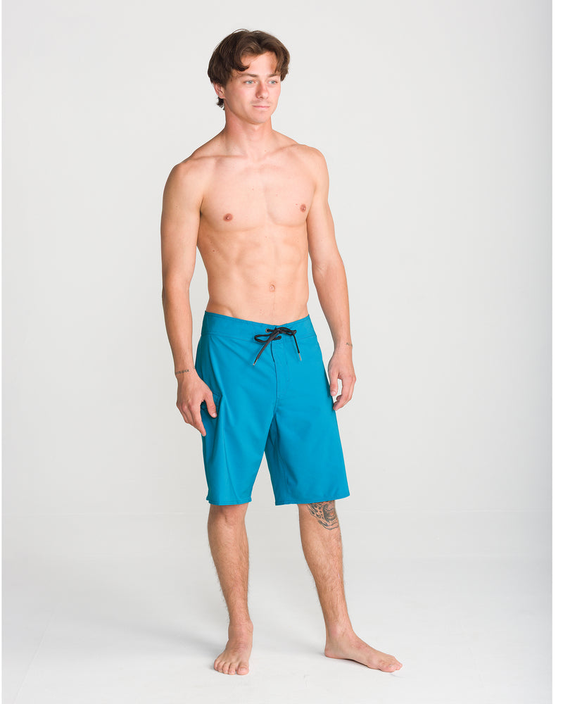 A young man with short brown hair stands barefoot on a plain background, wearing the Volcom Men’s Lido Solid Mod 20 boardshorts in a bright blue hue. The shorts feature 4-way stretch for ultimate comfort and mobility and are tied securely at the waist. He is shirtless, with a relaxed posture and a tattoo visible on his lower left leg.