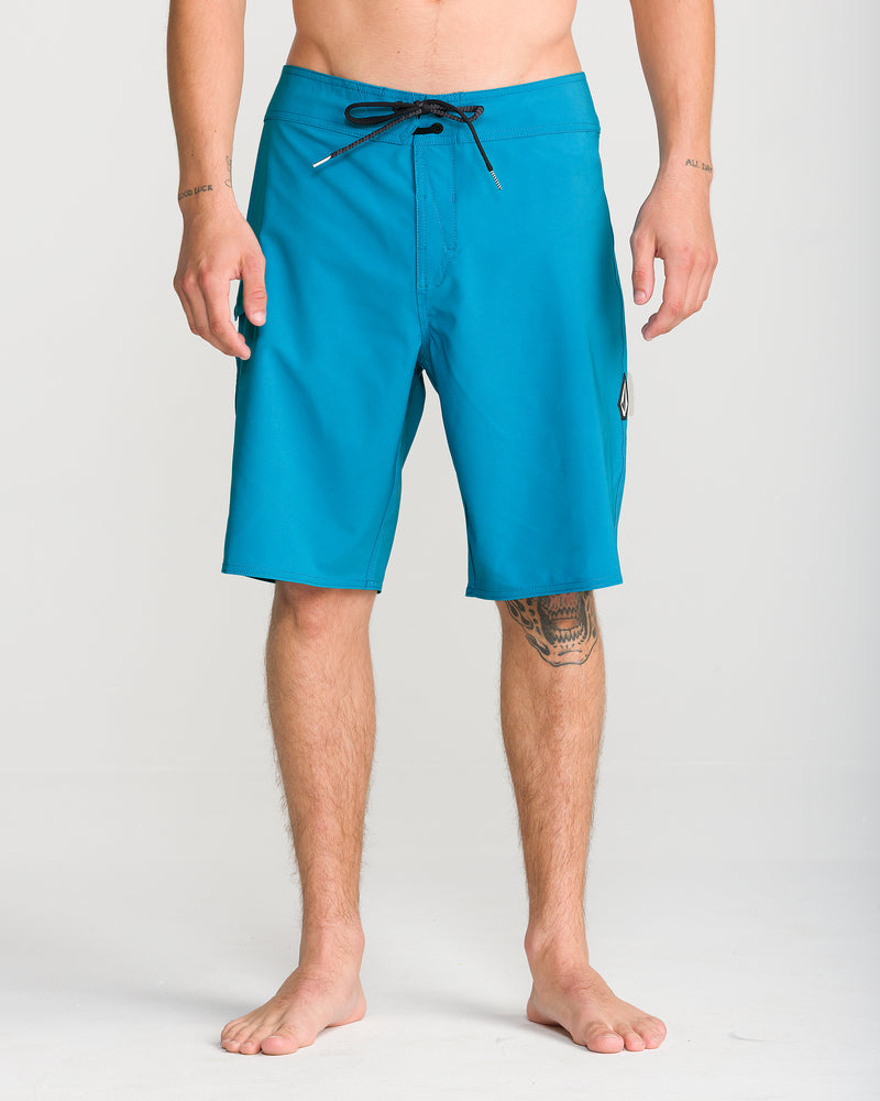 A person stands barefoot against a plain light background, wearing the Volcom Men’s Lido Solid Mod 20 boardshorts in bright blue. These shorts, which reach just above the knees, feature 4-way stretch for added comfort and mobility and are finished with a black drawstring.