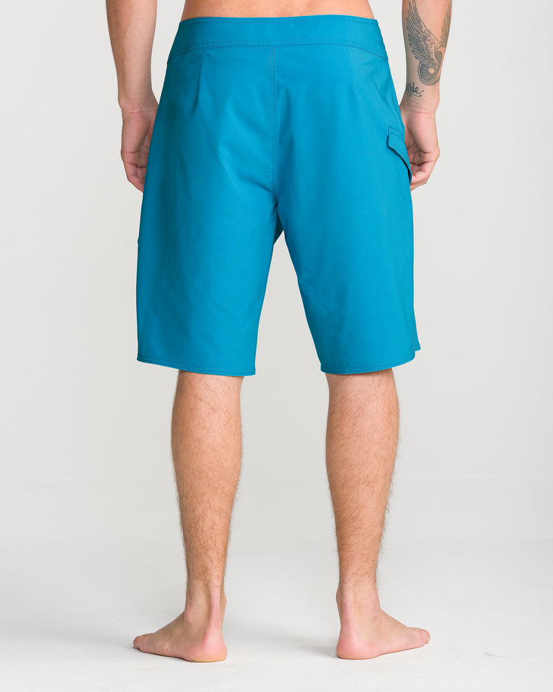 A person stands barefoot on a white floor, wearing Volcom Men’s Lido Solid Mod 20 blue boardshorts featuring Cinch Fly Technology. The image is taken from behind, showcasing the boardshorts and part of the person’s legs and arms.