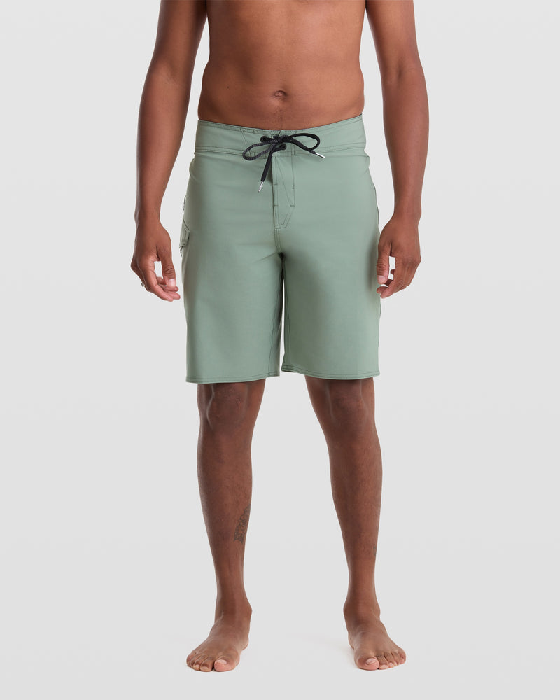 A barefoot man stands against a white background, wearing Volcom Men's Lido Solid 20" Trunks in light green with a black drawstring. Made from recycled polyester with 4-way stretch, these boardshorts offer comfort. He is shirtless and relaxed.