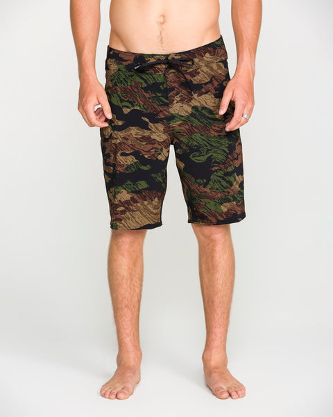 A person stands barefoot wearing Volcom Men's Lido Solid Mod board shorts, crafted from recycled fibers and featuring 4-way stretch for superior comfort. Their torso is visible from the waist down against a plain, light-colored background.