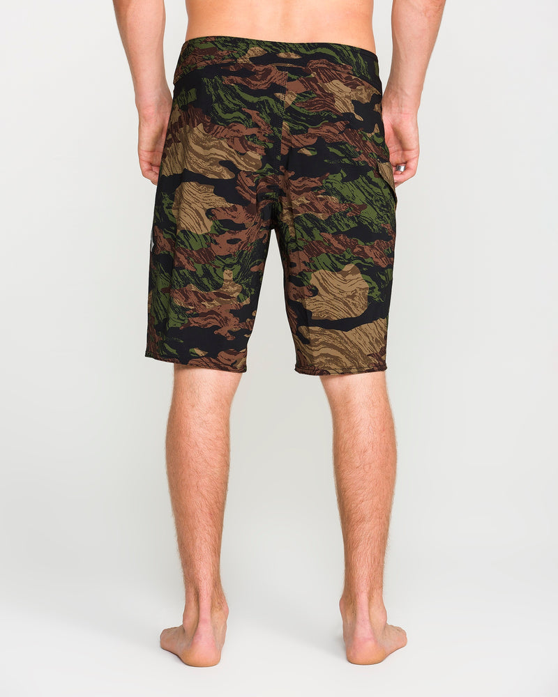 A person stands barefoot facing away, wearing green, brown, and tan knee-length camouflage shorts against a plain light background. The Volcom Men's Lido Solid Mod shorts feature a Cinch Fly and are made from 4-way stretch recycled fibers for comfort and mobility.