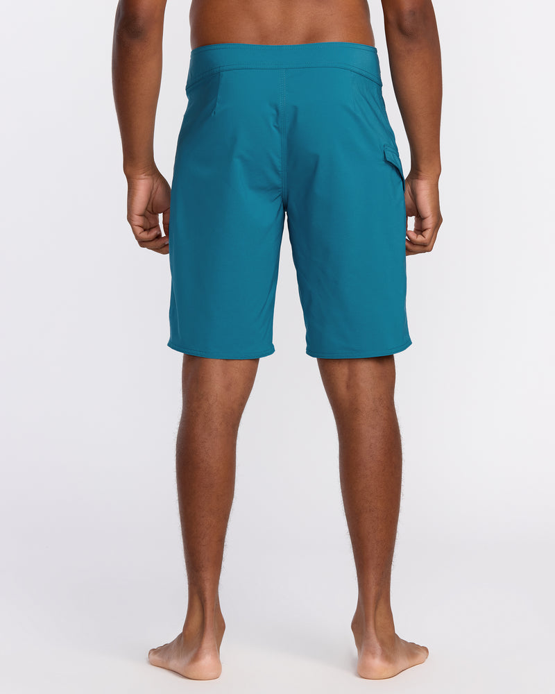 A man stands barefoot facing away from the camera, wearing Men's Lido Solid Mod Trunks—teal, knee-length boardshorts with pockets made from recycled polyester—against a plain white background.