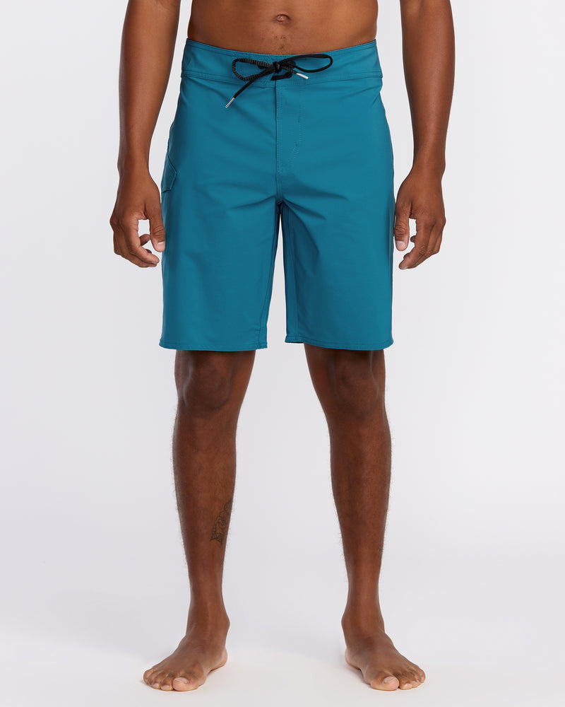 A person stands barefoot against a white background, wearing Men's Lido Solid Mod Trunks in teal with a black drawstring. Their arms rest at their sides, showing the trunks' knee-length fit and comfortable stretch fabric.