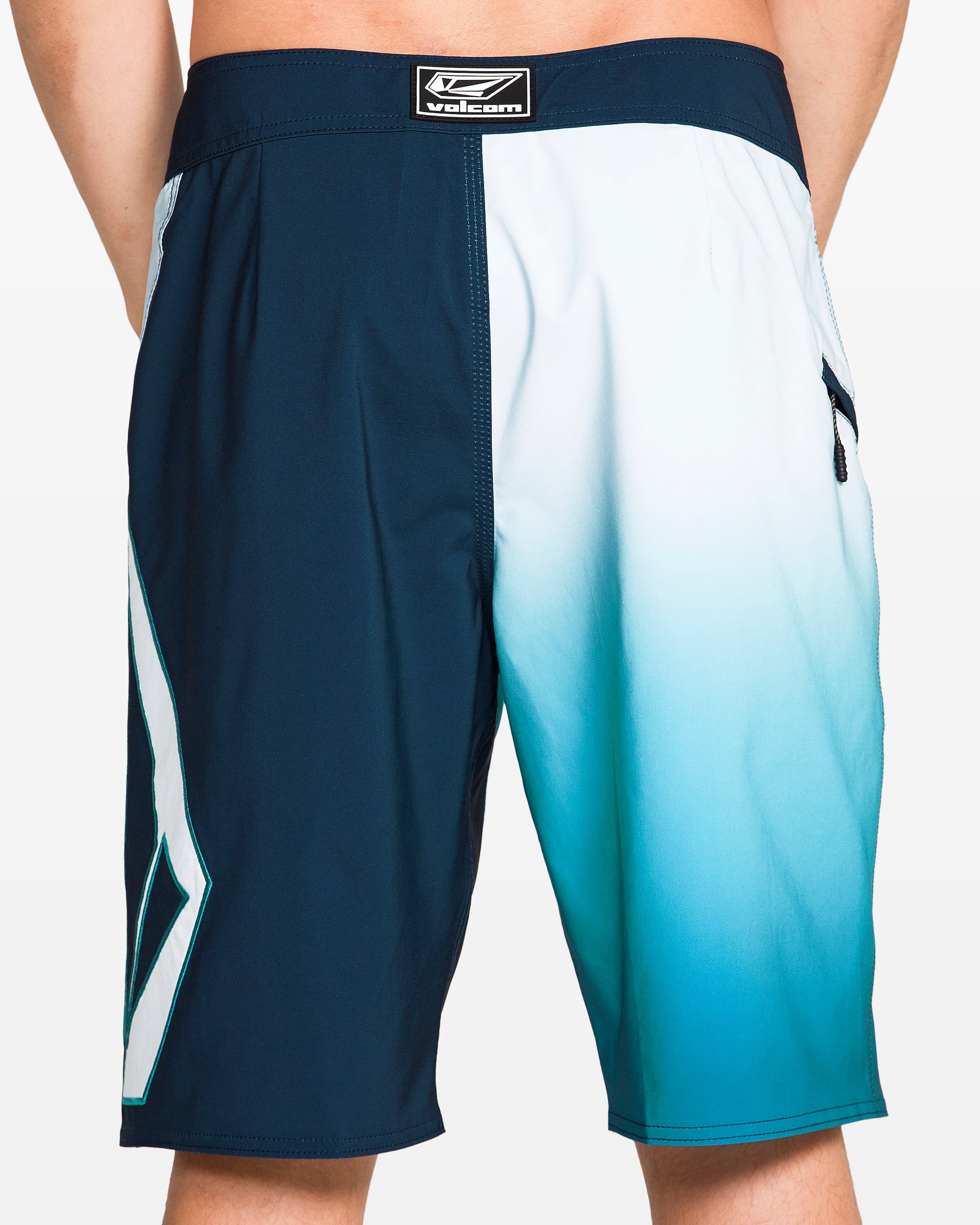 A person stands with hands behind their back wearing Volcom Men's Foster Mod 21" boardshorts featuring a blue and white ombre design, geometric pattern on one leg, Cinch Fly Technology, and a Velcro logo patch on the waistband.
