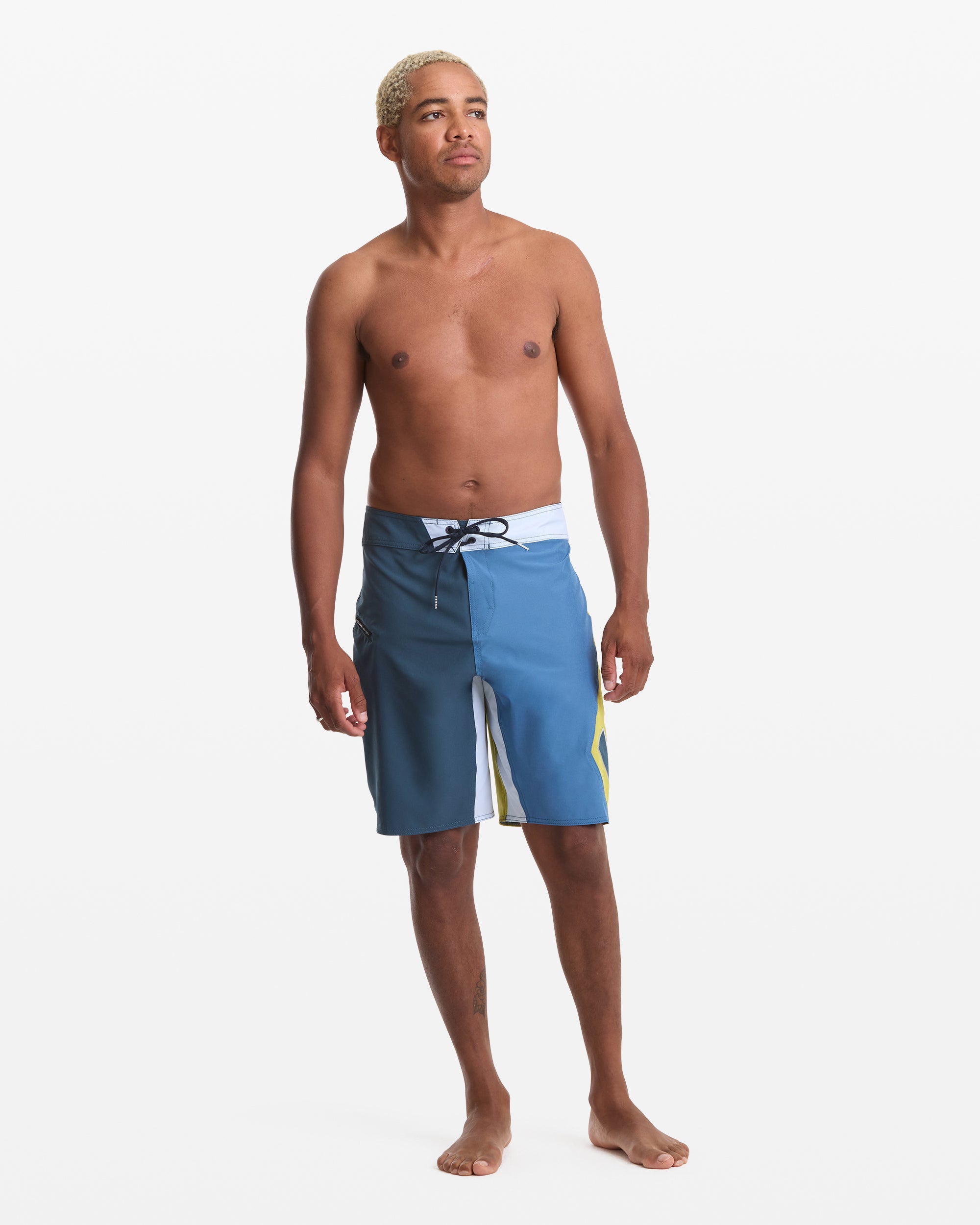 A barefoot man with short blond hair stands against a white background, wearing the Volcom Men's Foster Mod 21 blue board shorts with a black drawstring and white and yellow accents. He is shirtless and looking slightly to the side.