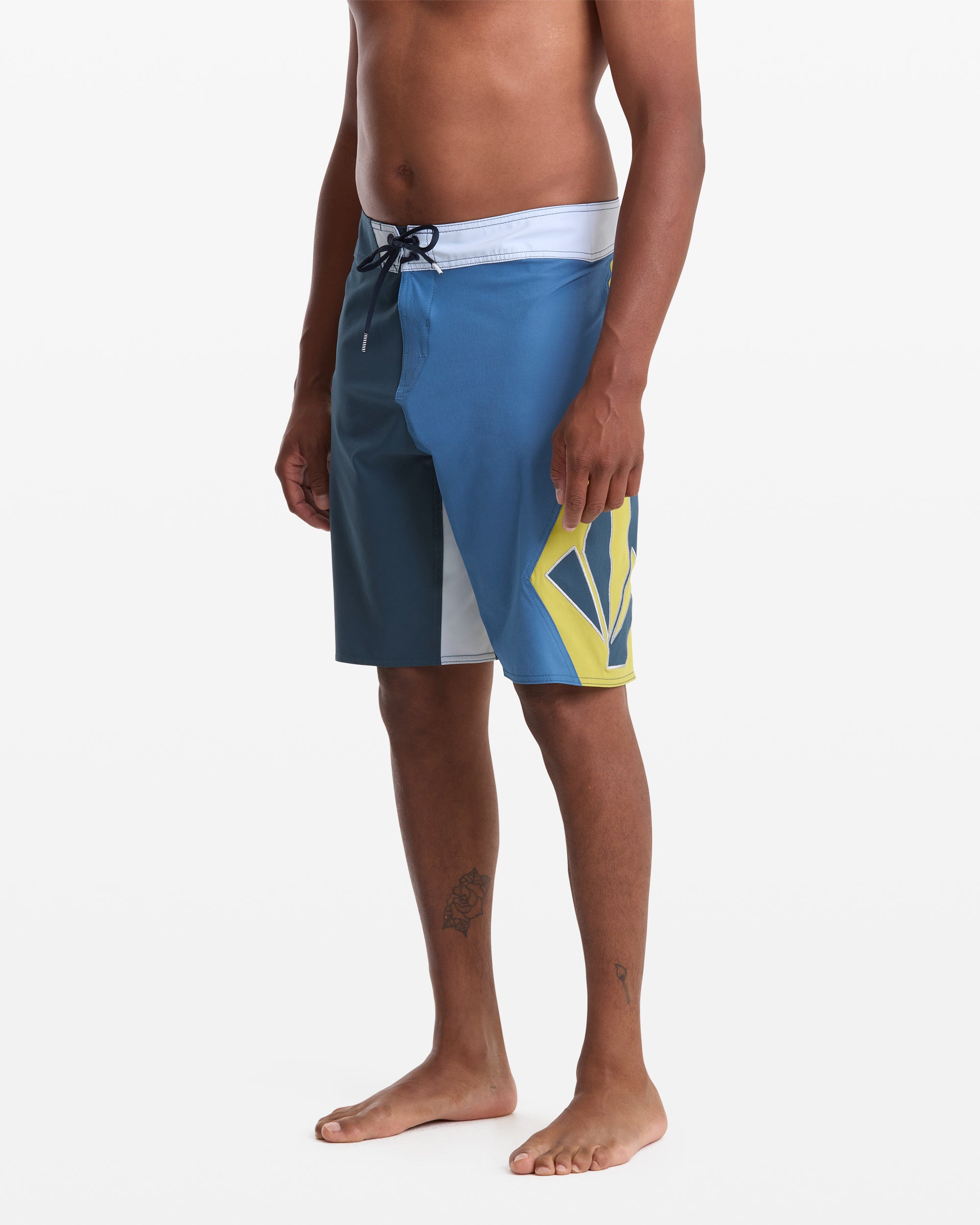 A man stands barefoot wearing Volcom Men's Foster Mod 21 board shorts with a white waistband, black drawstring, and bold yellow and navy geometric pattern on one leg. His upper body is not fully visible.