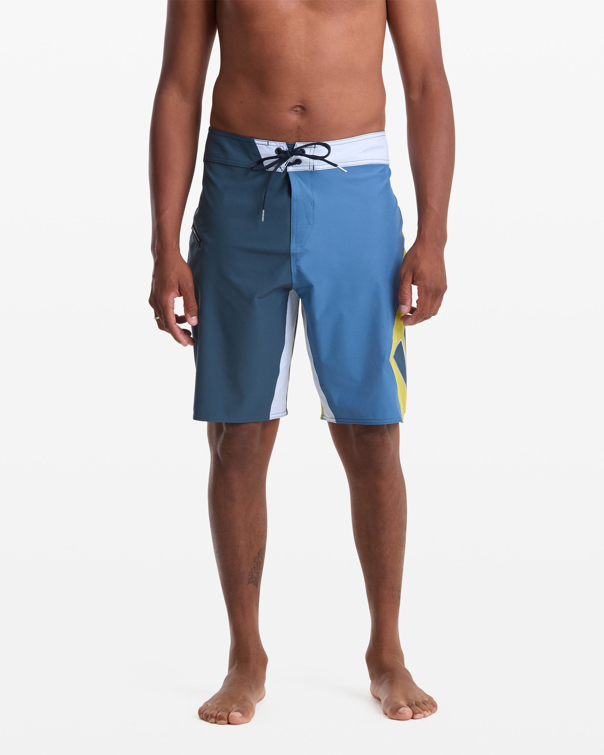A person models the Volcom Men's Foster Mod 21 boardshorts in blue and white with a yellow accent, featuring a drawstring waist and above-the-knee length, standing barefoot against a plain white background.