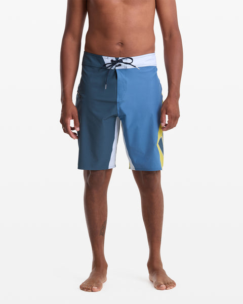 A person models the Volcom Men's Foster Mod 21 boardshorts in blue and white with a yellow accent, featuring a drawstring waist and above-the-knee length, standing barefoot against a plain white background.