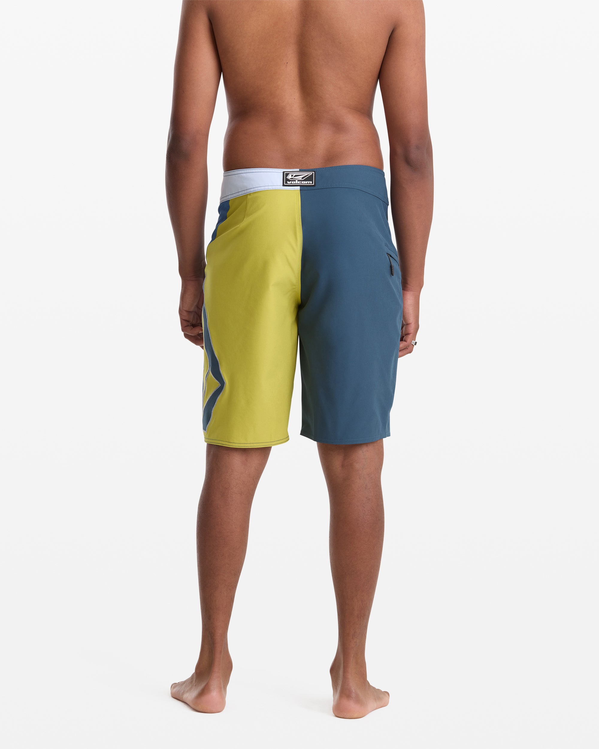 A man stands barefoot facing away from the camera, wearing Volcom Men's Foster Mod 21 two-tone board shorts—one yellow leg and one blue leg—against a plain white background.