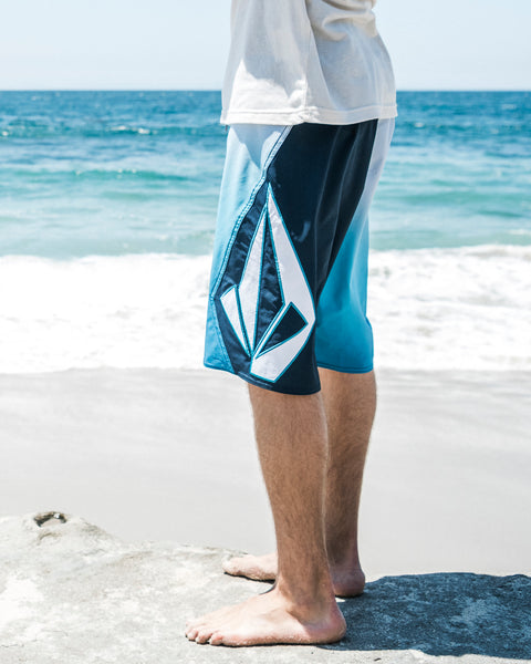 A person stands barefoot on the sand, wearing Volcom Men's Foster Mod boardshorts with a geometric design and a 21-inch outseam. Waves roll in behind them beneath a bright sky.
